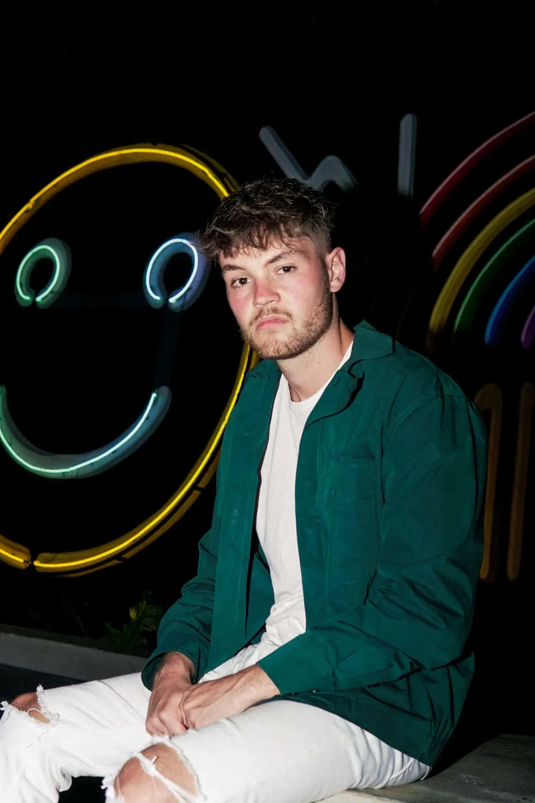 Young man in a green jacket seated against a colorful backdrop